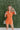 Frontal view of the Spur The Moment Denim Romper that features an orange denim material, a surplice neckline that snaps with a collar, half-length sleeves, a front zipper, pockets, pleating on the legs, and a 3" inseam.