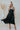 Frontal view of the Stealing Hearts Midi Dress that features a black colored silk material, a surplice neckline, a sleeveless design with thin adjustable straps, a pleated skirt, an open back with crossover straps and adjustable ties, and a midi length
