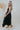 Side view of the Stealing Hearts Midi Dress that features a black colored silk material, a surplice neckline, a sleeveless design with thin adjustable straps, a pleated skirt, an open back with crossover straps and adjustable ties, and a midi length