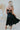 Back view of the Stealing Hearts Midi Dress that features a black colored silk material, a surplice neckline, a sleeveless design with thin adjustable straps, a pleated skirt, an open back with crossover straps and adjustable ties, and a midi length