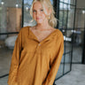 Frontal view of the Honey Honey Top that features a camel colored cotton material, a round neck, a 3/4 button-up front, a long sleeve, exposed hems, side slits, a high-low design, and a flowy fit.