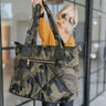 The Carryall Tote features a green and black camo print, a nylon material, two snap closure outside pockets, an outside zipper pocket, a zipper pocket and two open pockets within, an adjustable crossbody strap, two shoulder straps, and a zipper closure.