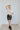 Full body front view of model wearing the Take It Back Skirt, that has a black faux-leather material, a high-rise fit, a faux belt with two buckles, faux pockets with stud detailing, a back zipper closure, and a mini length. Worn with shacket and white te