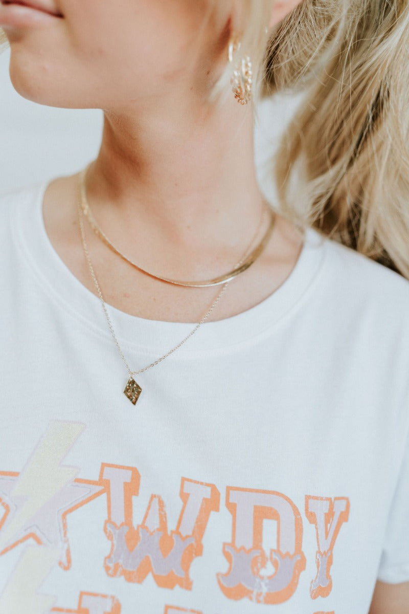 The Livin' The Dream Necklace is a gold, double layered necklace, featuring a thin chain with a diamond shaped pendant, a herringbone chain, and an adjustable link and clasp closure. 