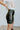 Side view of model wearing the Take It Back Skirt, that has a black faux-leather material, a high-rise fit, a faux belt with two buckles, faux pockets with stud detailing, a back zipper closure, and a mini length. 