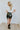 Full body back view of model wearing the Take It Back Skirt, that has a black faux-leather material, a high-rise fit, a faux belt with two buckles, faux pockets with stud detailing, a back zipper closure, and a mini length. 