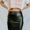 Front view of model wearing the Take It Back Skirt, that has a black faux-leather material, a high-rise fit, a faux belt with two buckles, faux pockets with stud detailing, a back zipper closure, and a mini length. 