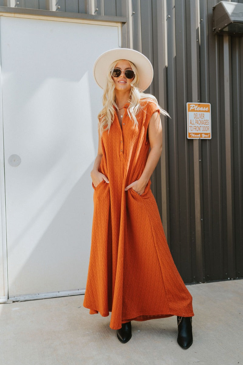 Frontal view of the Goodbye Summer Jumpsuit that features a rust colored knit material, a round neck, a 3/4 button-up front, a sleeveless design, side pockets, a wide leg, and an oversized fit.