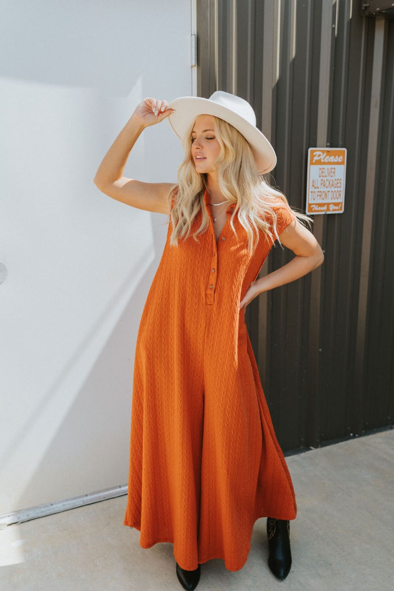Frontal view of the Goodbye Summer Jumpsuit that features a rust colored knit material, a round neck, a 3/4 button-up front, a sleeveless design, side pockets, a wide leg, and an oversized fit.