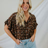 Frontal view of the Bare It All Top that features a brown chiffon material, a multi-colored floral print, a collar neck, a hidden button-up front, a short sleeve, a curved bottom hem side slits, and a high-low design.