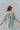 Back view of the Easy Breezy Top that features a green colored knit material, an open knit design, a V neckline, a long cuffed sleeve, side slits, and a high-low design.