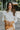 Frontal view of the Serenity Sweater that features a cream colored knit material, a high neckline, long cuffed sleeves, a surplice front with a tie closure, a side chest cut-out, and a thick bottom hem.