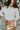 Frontal view of the Serenity Sweater that features a cream colored knit material, a high neckline, long cuffed sleeves, a surplice front with a tie closure, a side chest cut-out, and a thick bottom hem.