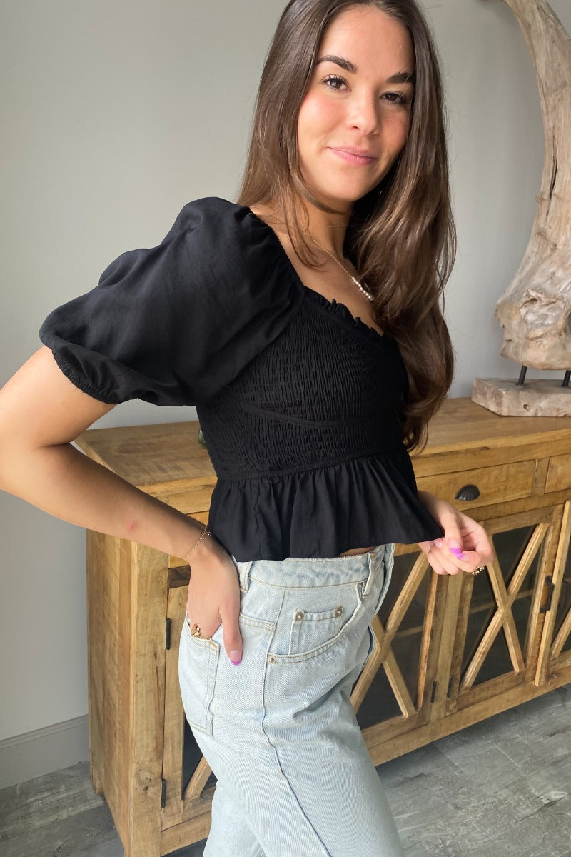 Side view of model wearing the Stand Alone Top which features a black material, a scoop neckline with a ruffle hem, a short puffy sleeve, a smocked front, a flowy bottom he, a cropped fit, a smocked back, and a tie back.