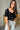 Front view of model wearing the Stand Alone Top which features a black material, a scoop neckline with a ruffle hem, a short puffy sleeve, a smocked front, a flowy bottom he, a cropped fit, a smocked back, and a tie back.