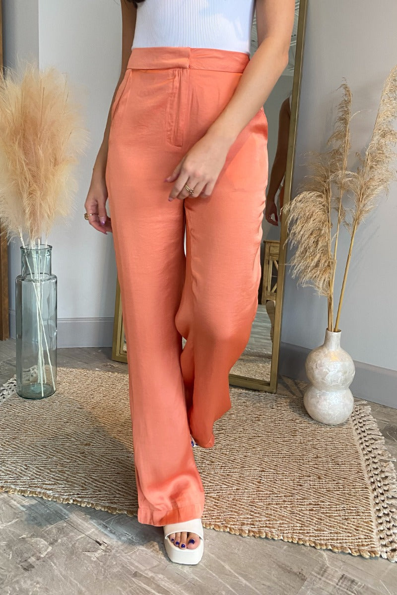 Front view of model wearing the Weekend Lover Pants which features orange satin fabric, two front pockets, a front zipper with hook closure, and flared legs.