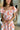 Close up view of model wearing the Let's Go Floral Dress which features white fabric with an orange, purple, and tan floral design, a mini length hem, a white lining, a smocked chest, a square neckline, and short sleeves with ruffle trim.
