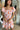 Front view of model wearing the Let's Go Floral Dress which features white fabric with an orange, purple, and tan floral design, a mini length hem, a white lining, a smocked chest, a square neckline, and short sleeves with ruffle trim.