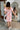Full body/back view of model wearing the Let's Go Floral Dress which features white fabric with an orange, purple, and tan floral design, a mini length hem, a white lining, a smocked chest, a square neckline, and short sleeves with ruffle trim.