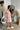 Side view of model wearing the Let's Go Floral Dress which features white fabric with an orange, purple, and tan floral design, a mini length hem, a white lining, a smocked chest, a square neckline, and short sleeves with ruffle trim.