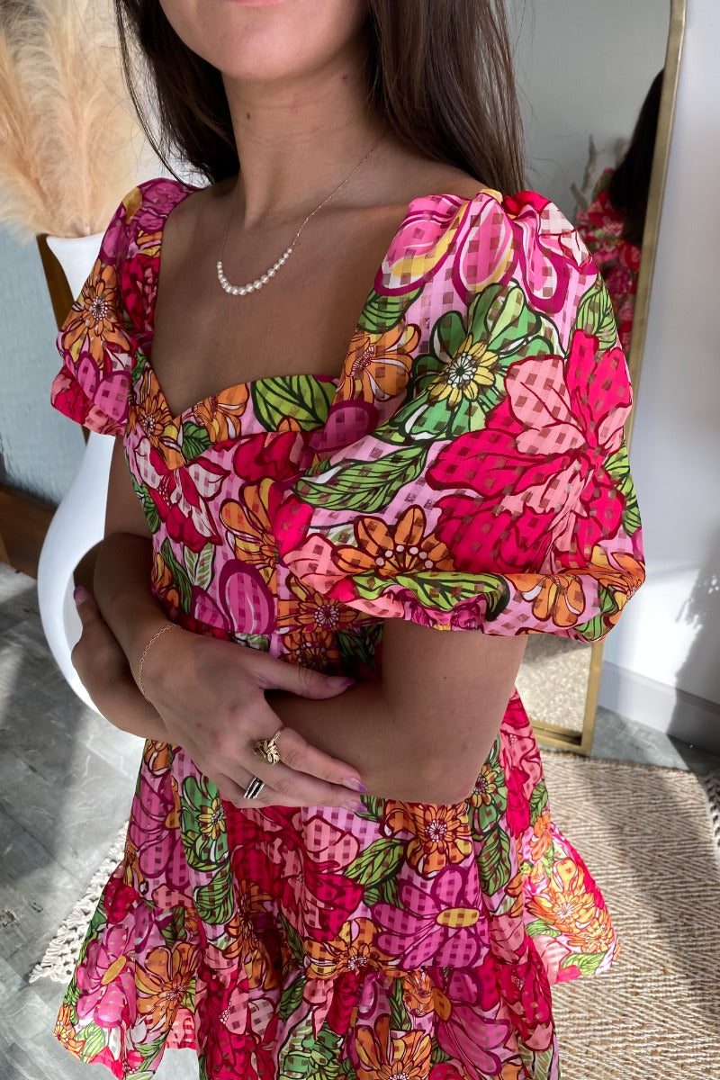 Close up/side view of model wearing the Best Memory Floral Dress which features hot pink, light pink, orange, yellow green and white fabric with a floral and gingham pattern, ruffle trim, a mini length hem, pink lining, a sweetheart neckline, a smocked ba