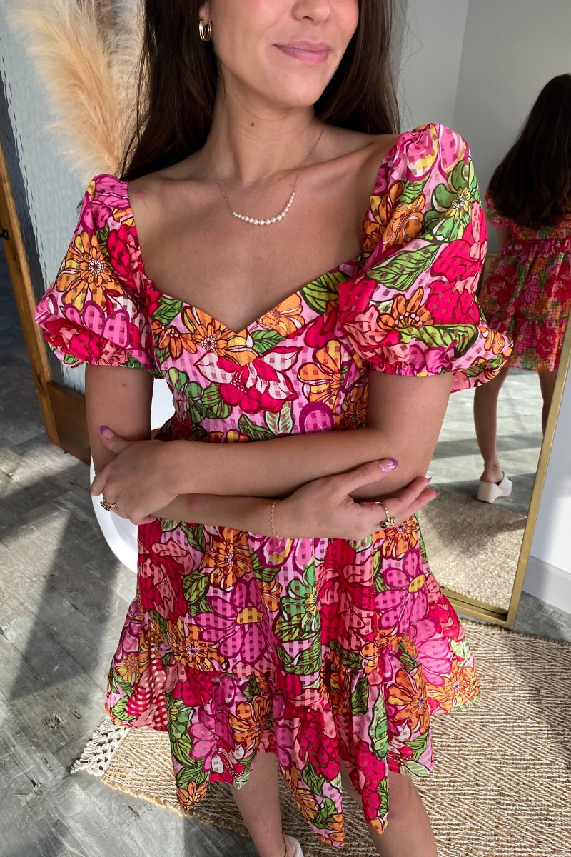 Close up view of model wearing the Best Memory Floral Dress which features hot pink, light pink, orange, yellow green and white fabric with a floral and gingham pattern, ruffle trim, a mini length hem, pink lining, a sweetheart neckline, a smocked back, a