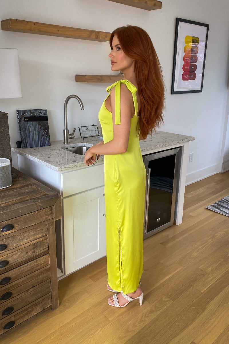Side view of model wearing the Shine Bright Dress which features neon lime satin fabric with a lining, a side slit, a scooped neckline, tie spaghetti straps, and a side zipper and hook closure.