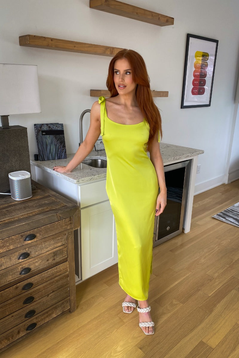 Full body view of model wearing the Shine Bright Dress which features neon lime satin fabric with a lining, a side slit, a scooped neckline, tie spaghetti straps, and a side zipper and hook closure.