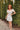 Full body view of model wearing the Loving Heart Dress which features white sheer fabric with a cream embroidered floral pattern, white lining, a cutout body design, a mini-length hem, ruffle details, an open back with a tie, a ruched sweetheart neckline 