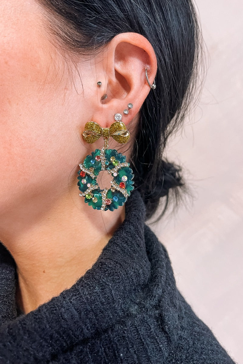 Side view of model wearing the Genevieve Green Wreath Earrings that have dangle, green christmas wreaths with multi color stones and clear stones alinked with a gold bow.