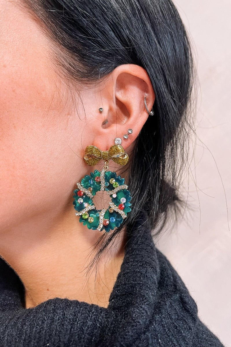 Side view of model wearing the Genevieve Green Wreath Earrings that have dangle, green christmas wreaths with multi color stones and clear stones linked with a gold bow.
