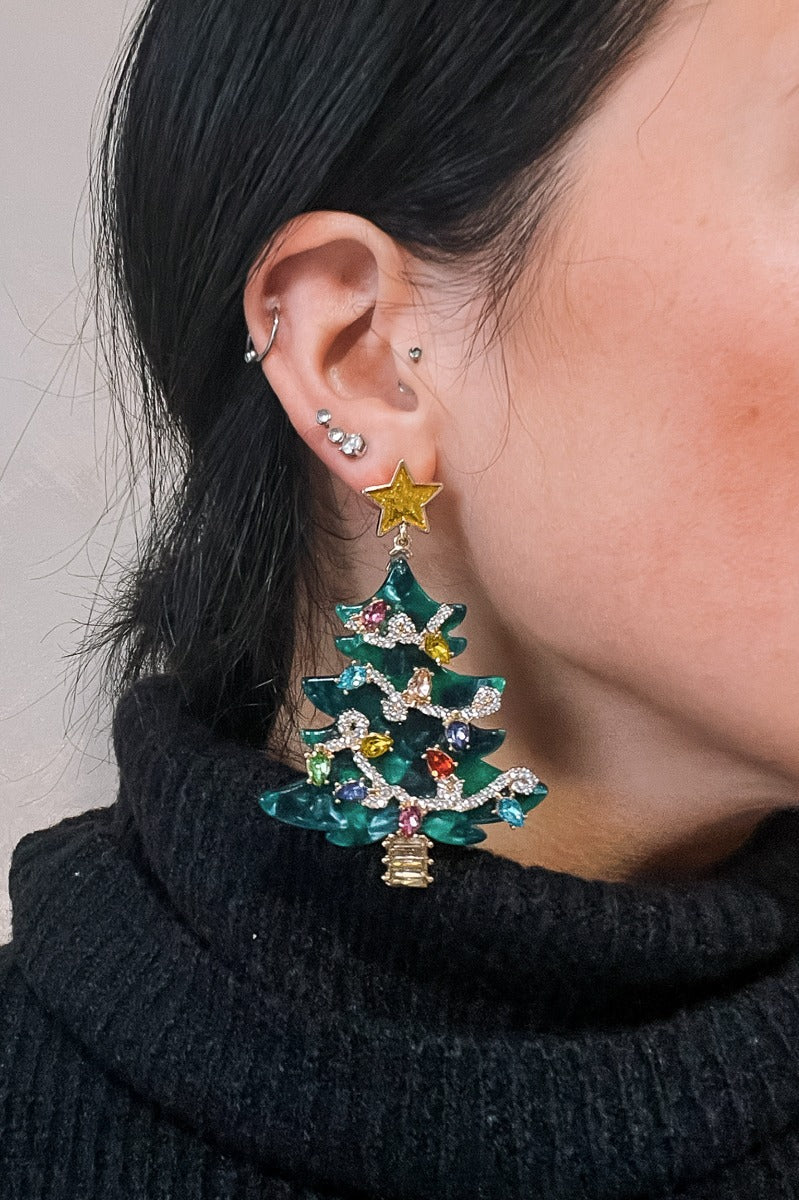 Close up of model wearing the Katherine Christmas Tree Earrings that have green christmas trees with multi color stones as lights and clear stones as strands linked with a gold star and yellow stone tree stump.