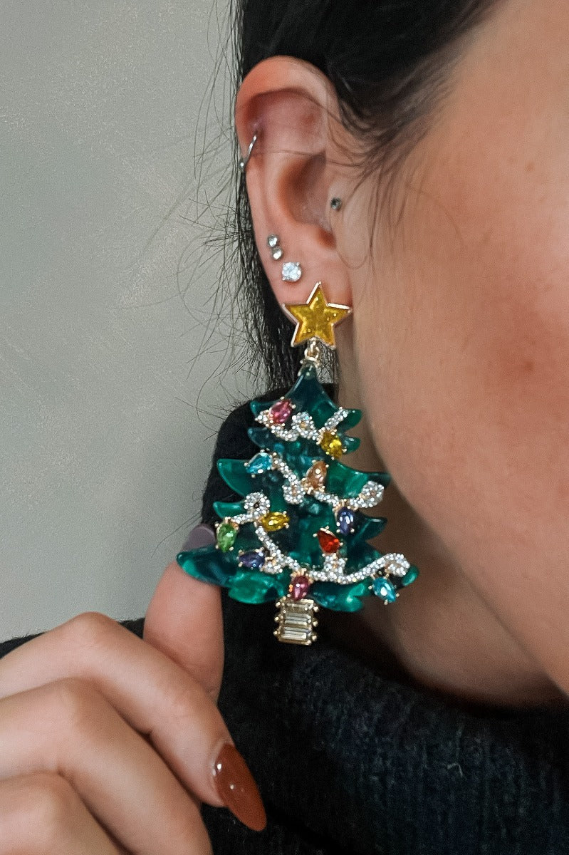 Close up of model wearing the Katherine Christmas Tree Earrings that have green christmas trees with multi color stones as lights and clear stones as strands linked with a gold star and yellow stone tree stump.