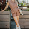 Full body front view of model wearing the Desert Dreams Romper that has brown fabric, pockets, buttons, an adjustable belt, an elastic waist, a collared neck, and a sleeveless design