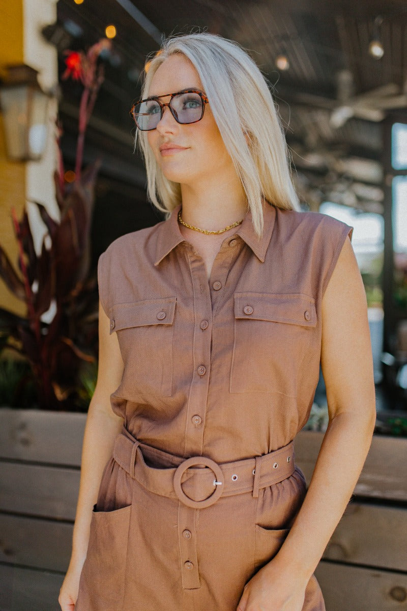 Upper front view of model wearing the Desert Dreams Romper that has brown fabric, pockets, buttons, an adjustable belt, an elastic waist, a collared neck, and a sleeveless design