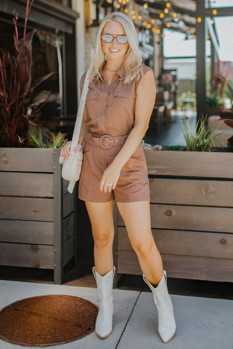 Full body front view of model wearing the Desert Dreams Romper that has brown fabric, pockets, buttons, an adjustable belt, an elastic waist, a collared neck, and a sleeveless design