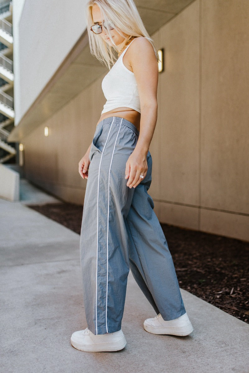 Full body side view of model wearing the Ready To Go Pants that have grey lightweight fabric, white stripe side details, an elastic waistband, two front pockets and straight leg pants