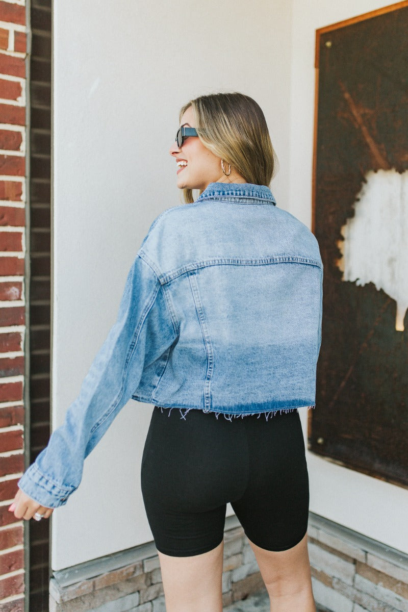 Back view of model wearing the Over it Cropped Denim Jacket that has blue denim fabric, a cropped waist with a raw hem, a button up front, pockets, a collared neck, and long sleeves.