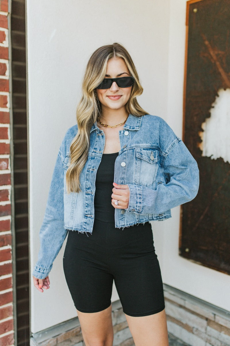 Front view of model wearing the Over it Cropped Denim Jacket that has blue denim fabric, a cropped waist with a raw hem, a button up front, pockets, a collared neck, and long sleeves.
