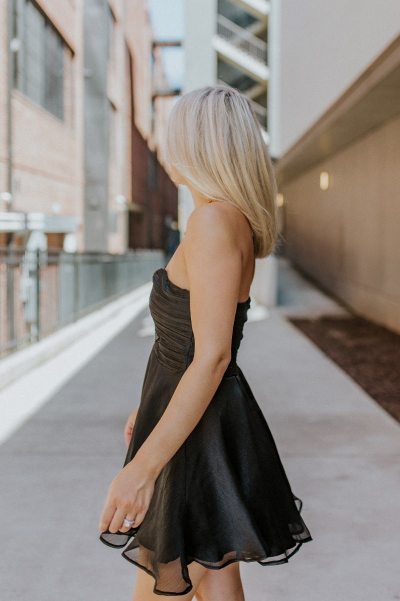 Side view of model wearing the Love Of Mine Dress that has black sheer fabric, mini length, black lining, a strapless sweetheart neckline with pleating, and a smocked back.