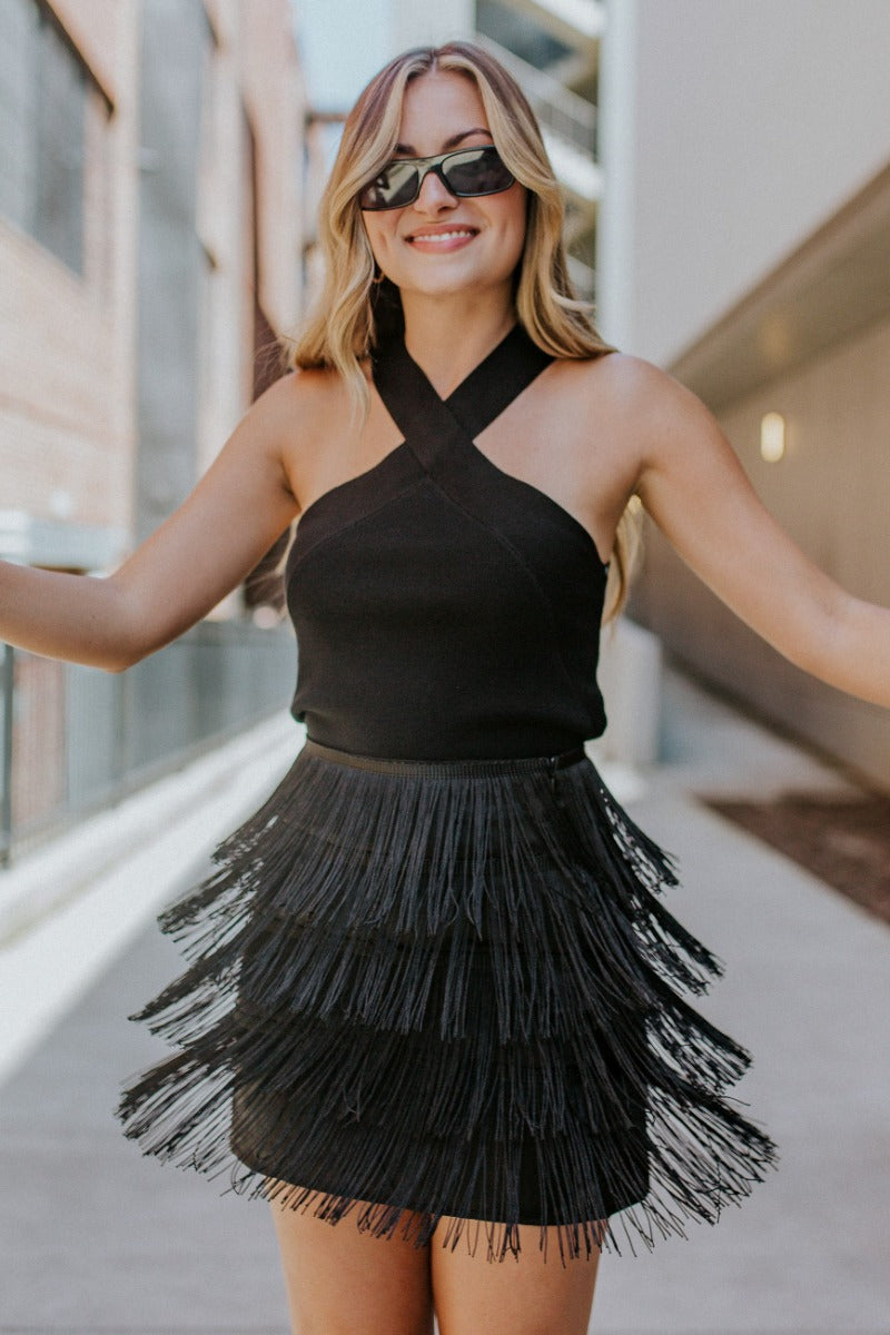 Front view of model wearing the Girls Want To Have Fun Fringe Skirt that has black satin fabric, a black fringe layered design, and black lining.