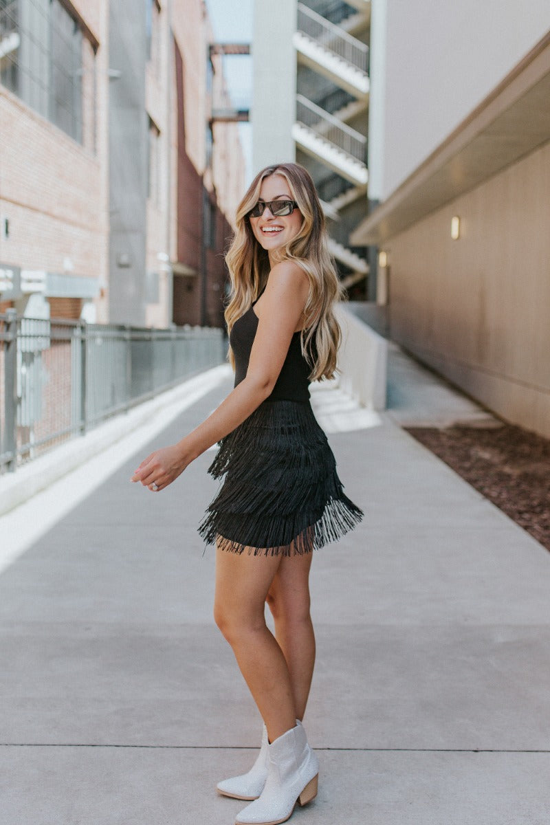 Full body side view of model wearing the Girls Want To Have Fun Fringe Skirt that has black satin fabric, a black fringe layered design, and black lining.