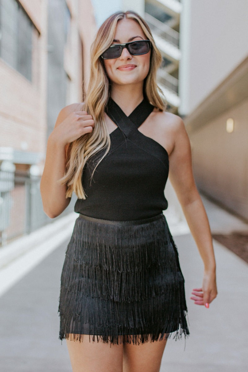 Front view of model wearing the Girls Want To Have Fun Fringe Skirt that has black satin fabric, a black fringe layered design, and black lining.