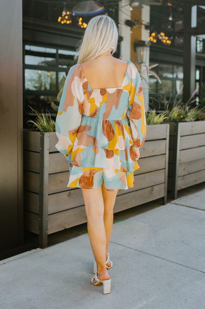 Back view of model wearing the In The Groove Romper that has sheer fabric with a mocha brown, taupe, pink, aqua, cream and mustard marbled pattern, a two-tiered ruffle skirt, a smocked upper, a square neck and long sleeves.