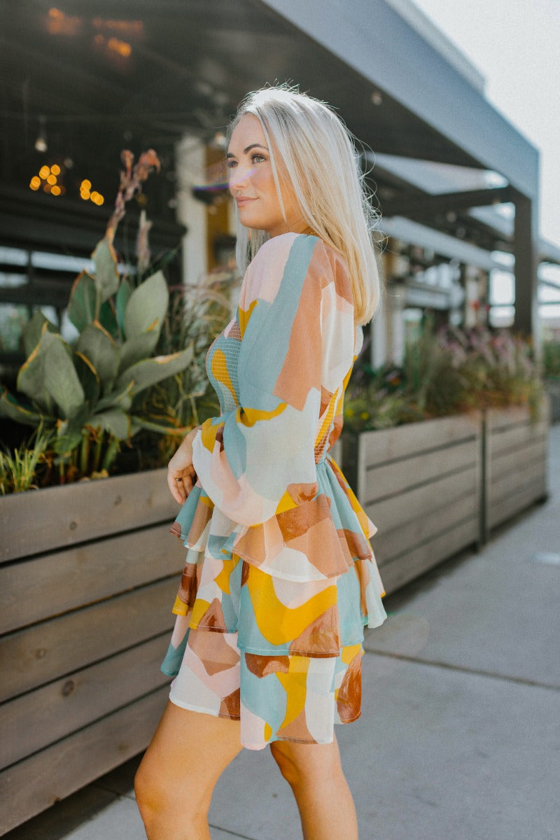 Side view of model wearing the In The Groove Romper that has sheer fabric with a mocha brown, taupe, pink, aqua, cream and mustard marbled pattern, a two-tiered ruffle skirt, a smocked upper, a square neck and long sleeves.