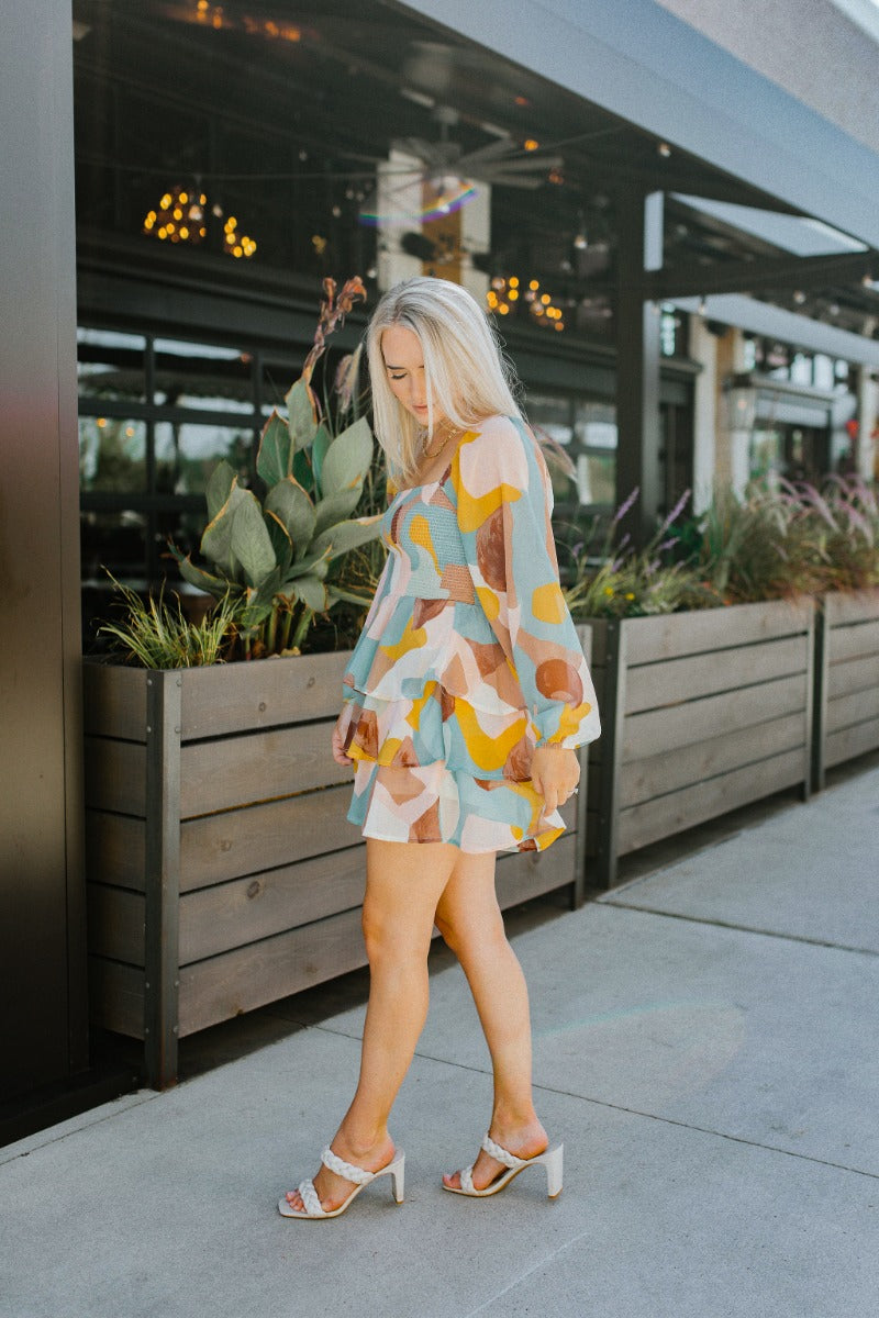 Full body side view of model wearing the In The Groove Romper that has sheer fabric with a mocha brown, taupe, pink, aqua, cream and mustard marbled pattern, a two-tiered ruffle skirt, a smocked upper, a square neck and long sleeves.