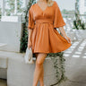 Full front view of model wearing the Spice Of Life Dress that has rust satin fabric, mini length, rust lining, an elastic waistband in the back, a tie around the waist, a v-neckline, short flare sleeves, and a back key hole with a button closure.