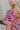 Close up side view of model wearing the Falling For You Floral Dress which features berry pink, purple, green and white sheer fabric, floral print, berry pink lining, front slit, ruffle layered hem, sweetheart neckline, short puff sleeve with ruffle hem a