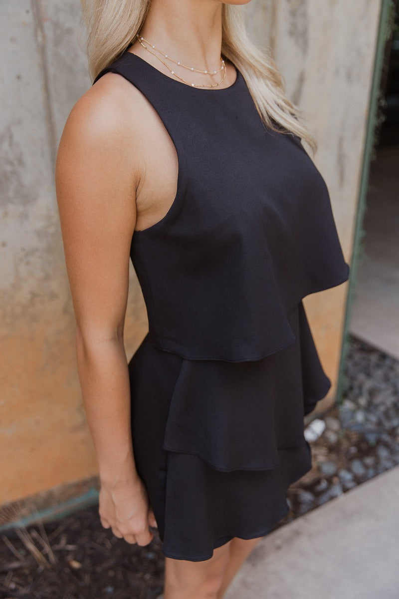 Side view of model wearing the All Over Again Romper which features black lightweight fabric, shorts lining, tiered ruffle design, round neckline, sleeveless and monochromatic back zipper with hook closure.