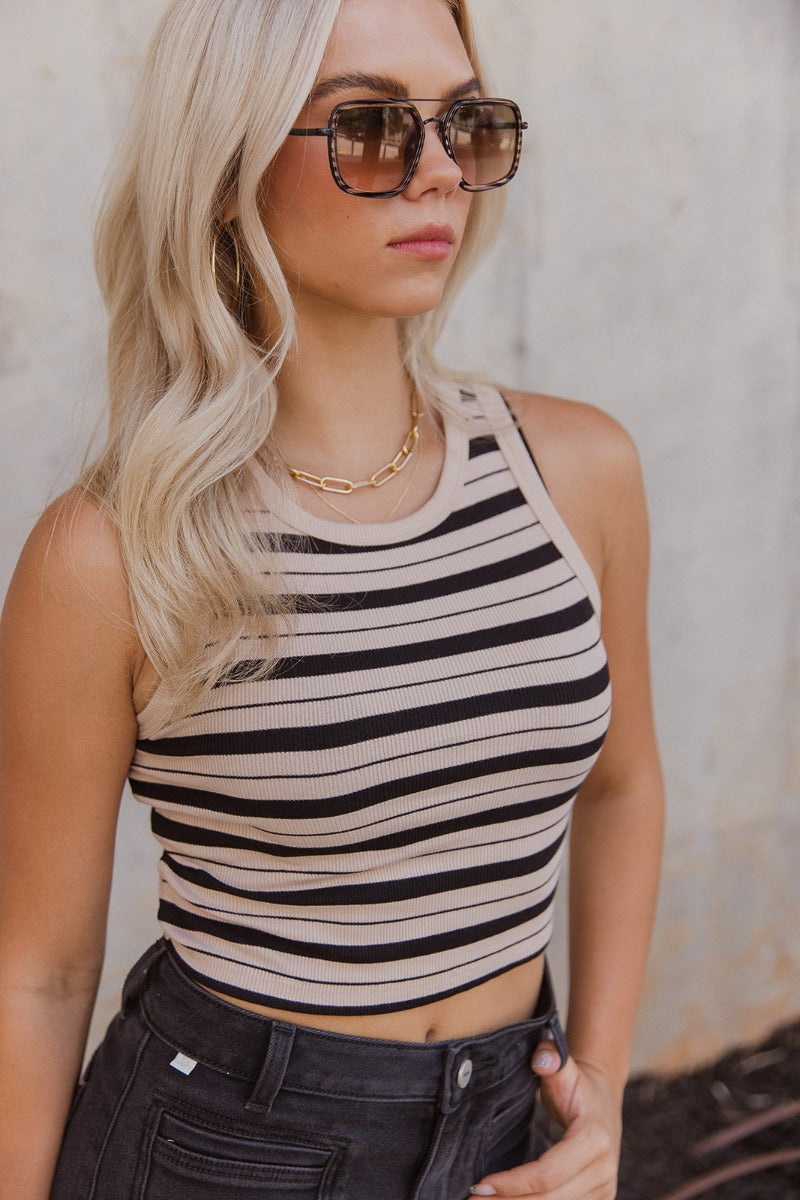 Close front view of model wearing the Keep It Simple Striped Tank that has ribbed fabric with a black and tan stripe pattern, a cropped waist, a round neckline and a sleeveless design